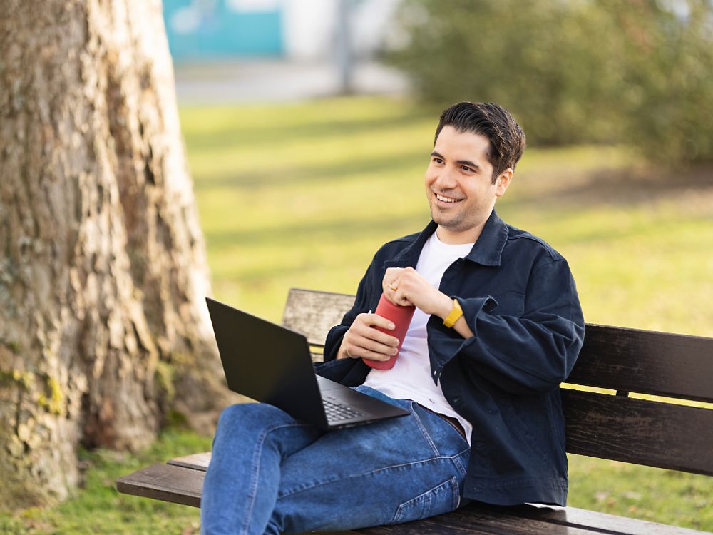 Un empleado de Henkel se sienta en un banco en un parque y trabaja en su computadora portátil. 