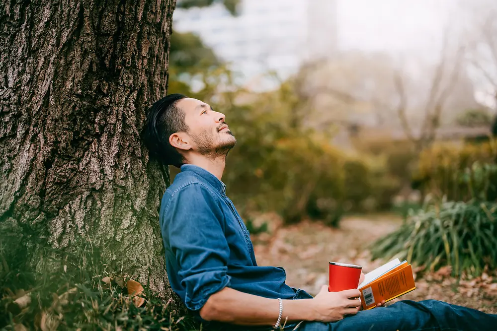 Un hombre junto a un árbol leyendo un libro.