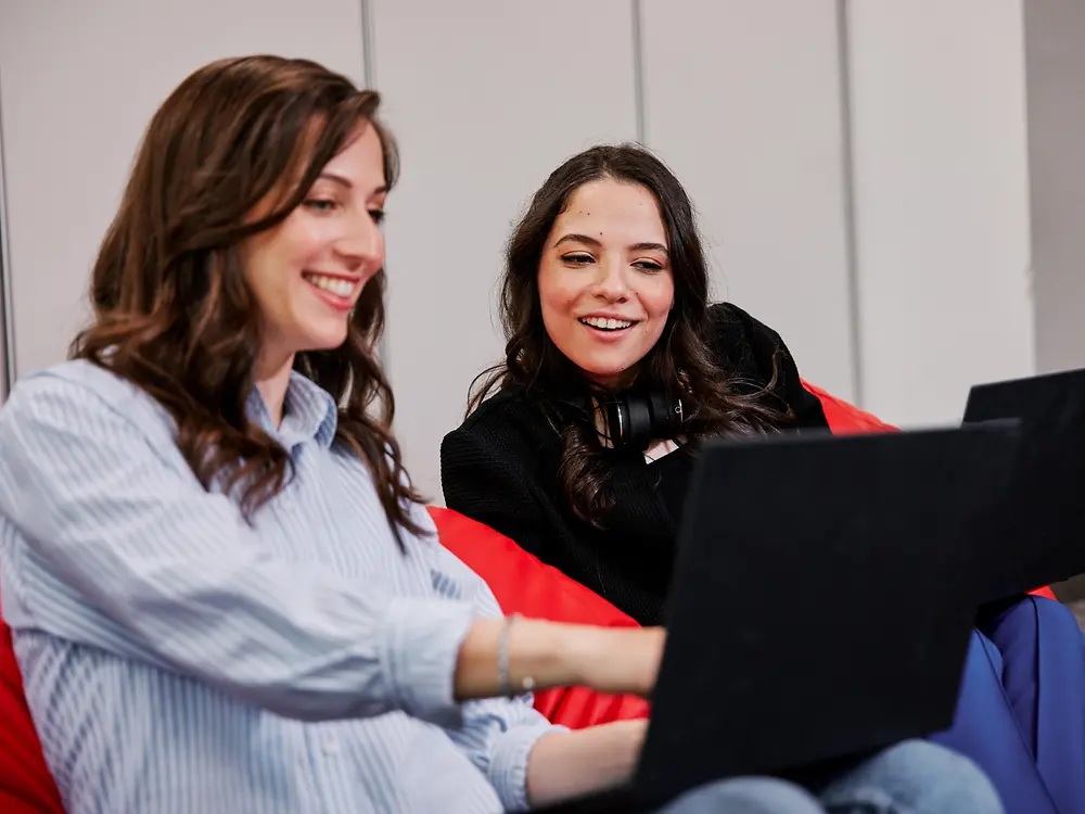 Dos personas sentadas en coloridos pufs en una oficina moderna, cada una usando una computadora portátil. Parece que están hablando de los beneficios para los empleados de Henkel mientras trabajan en colaboración.
