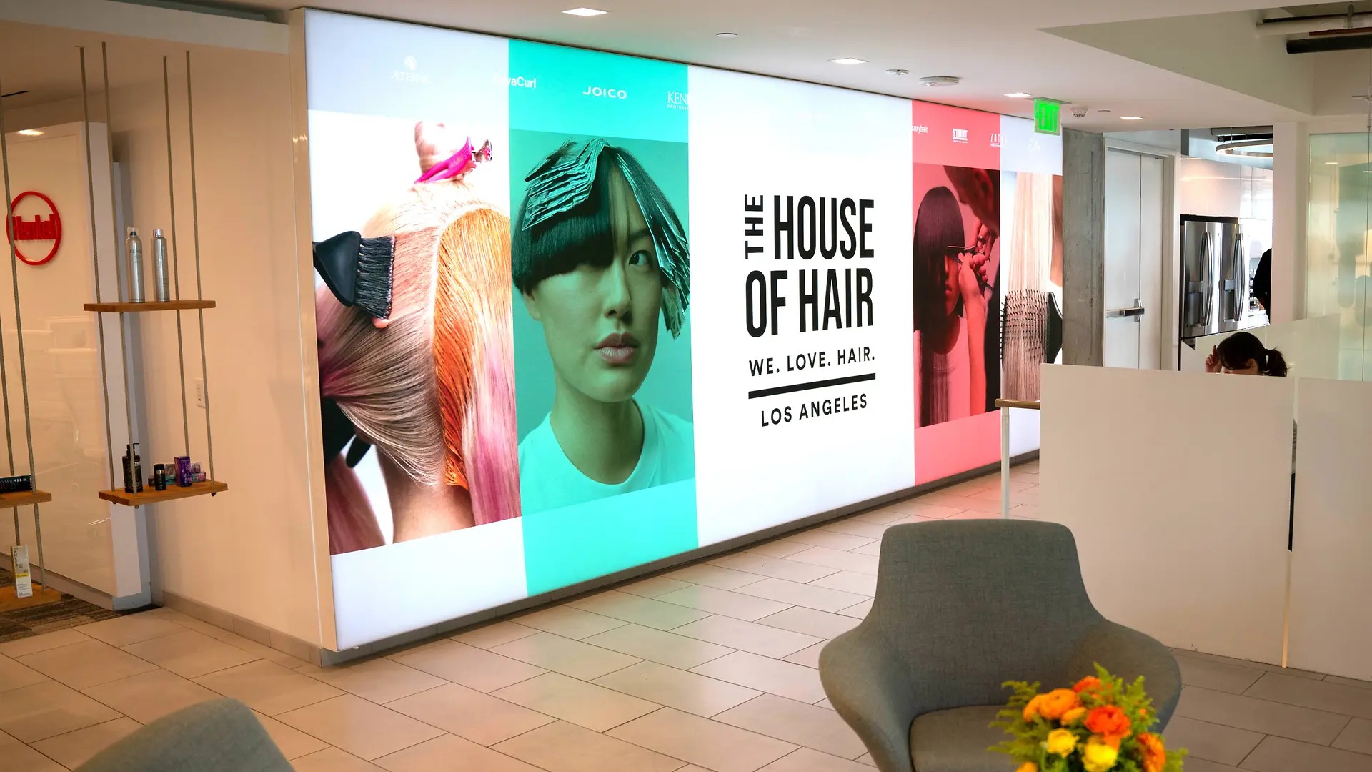 Un vestíbulo moderno con una luminosa exposición mural compuesta por grandes paneles con coloridos gráficos relacionados con el cabello y el texto "THE HOUSE OF HAIR – WE. LOVE. HAIR. – LOS ANGELES". El espacio incluye sillones de color gris claro, una pequeña mesa con un vibrante arreglo floral, así como un diseño limpio y minimalista con suelo de baldosas y tabiques blancos. 