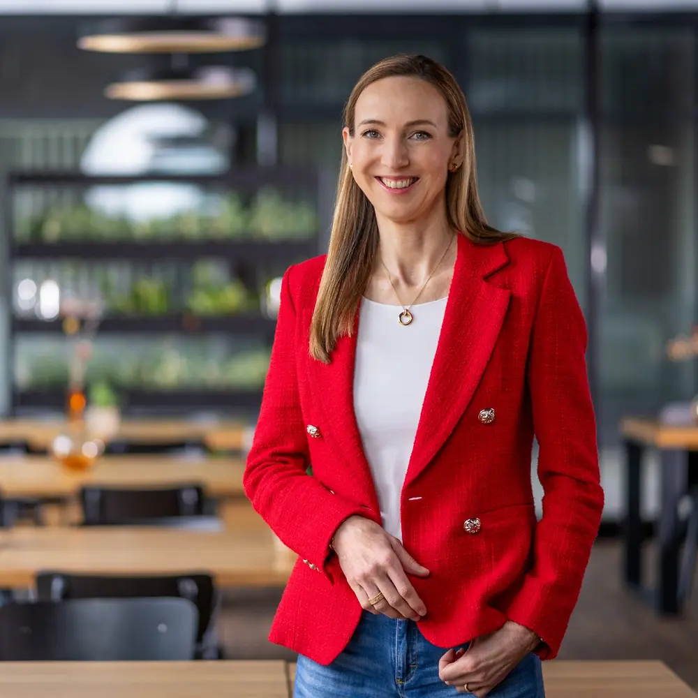 La Dra. Simone Bagel-Trah vistiendo un blazer rojo brillante y una blusa blanca, de pie en un espacio moderno tipo cafetería, con mesas de madera y jarrones decorativos al fondo.