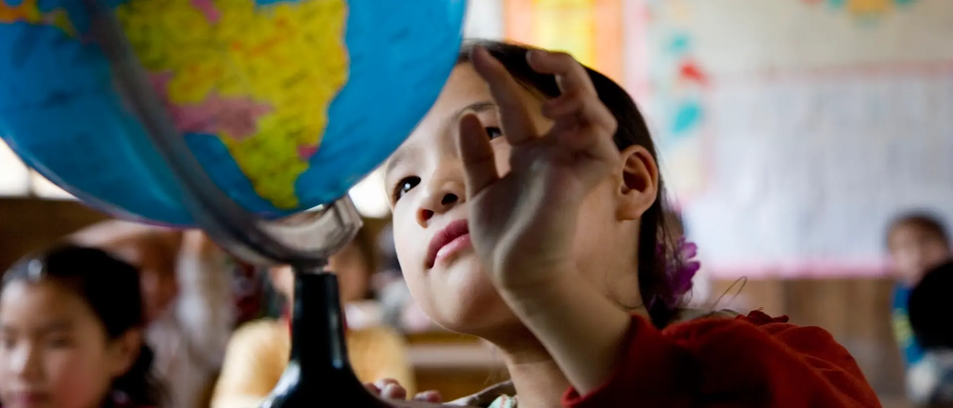 Un niño en un aula se inclina hacia un gran globo terráqueo colocado sobre un escritorio, con mapas y carteles coloridos visibles en las paredes del fondo.
