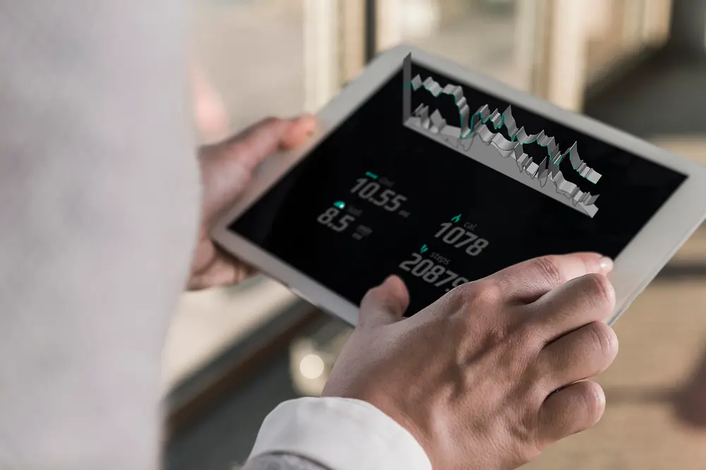 Persona sosteniendo una tablet que muestra gráficos digitales y datos numéricos, vista por encima del hombro en un entorno interior iluminado.