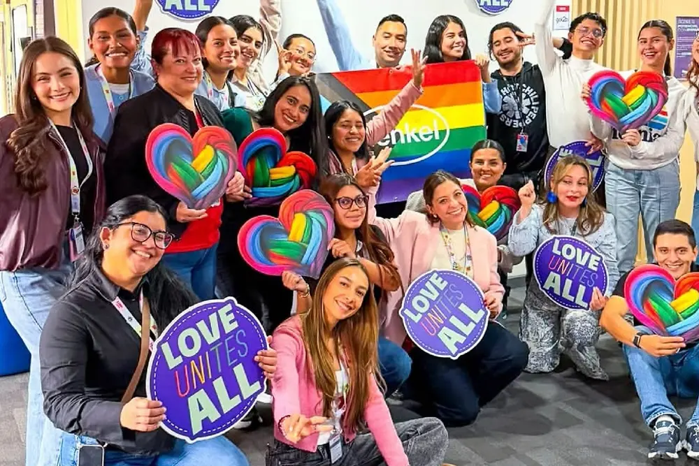 Foto grupal de aproximadamente 18 empleados reunidos en una oficina, sosteniendo coloridos carteles con temática de arcoíris, accesorios en forma de corazón y una bandera de arcoíris. Varios letreros muestran el texto “EL AMOR UNE A TODOS”. El grupo está dispuesto de pie y sentado en el piso alfombrado de una oficina, con escritorios, sillas y accesorios de oficina visibles al fondo. En la esquina inferior izquierda de la imagen aparece una etiqueta de ubicación que dice “Colombia”.