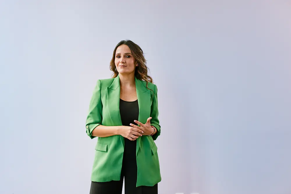 Retrato de una persona de pie sobre un fondo claro, con una chaqueta verde sobre una blusa oscura.